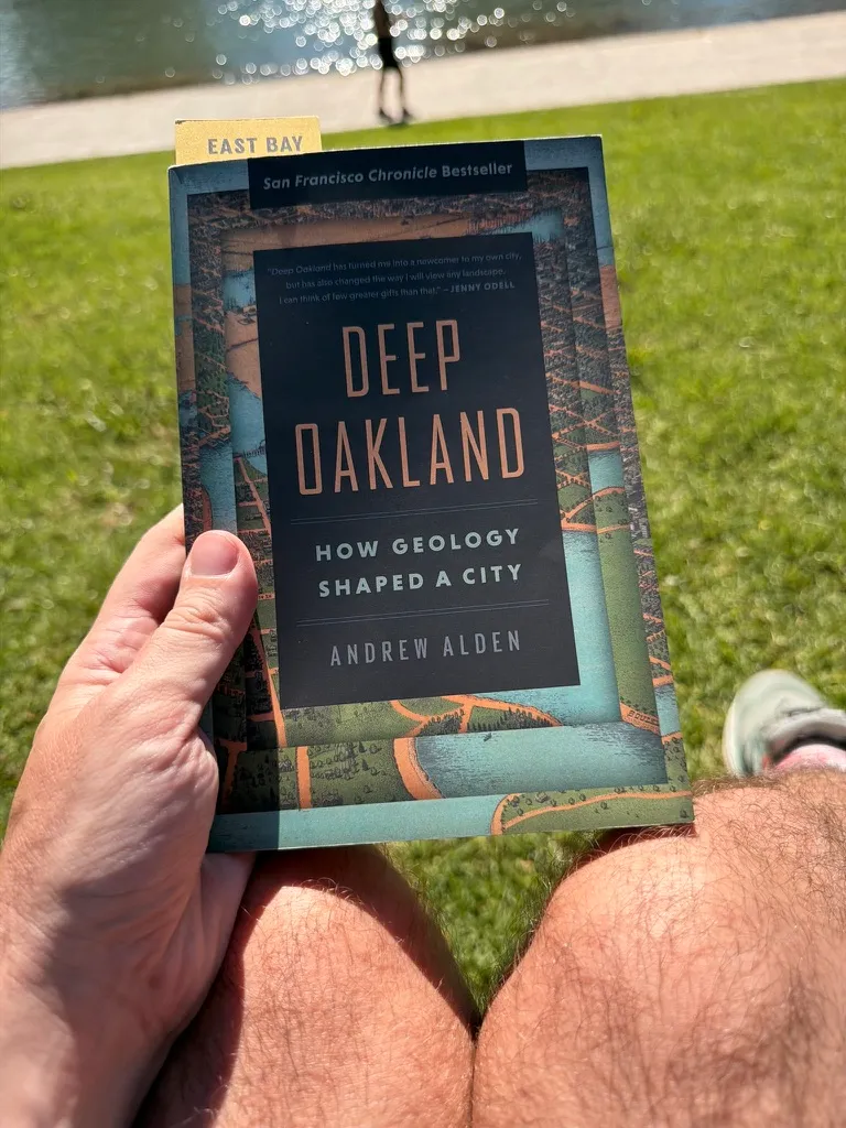 Keith holding a copy of Deep Oakland on his knees in the grass by the water