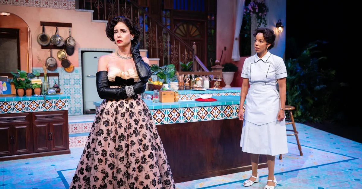 Production photo from The Cook: a woman in a ball gown and long black gloves and a woman in a white cook's uniform in a tiled Havana kitchen