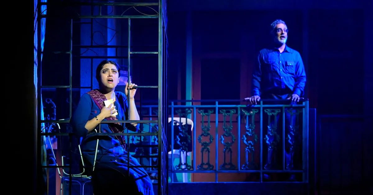 Production photo from The Lunchbox: a young woman holding a note on a Mumbai balcony while an older man looks on from the balcony across, both lit in blue